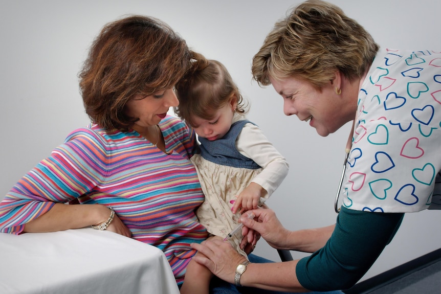 A female nurse in scrubs decorated with lovehearts administers a vaccine to a young toddler girl, being c