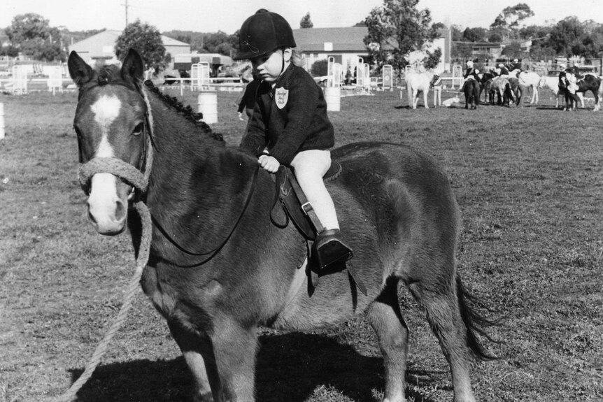 A black and white image of a little girl with jodhpurs, jacket and helmet, riding a pony.