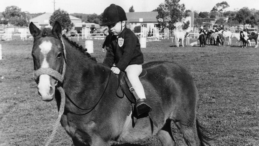 A black and white image of a little girl with jodhpurs, jacket and helmet, riding a pony.