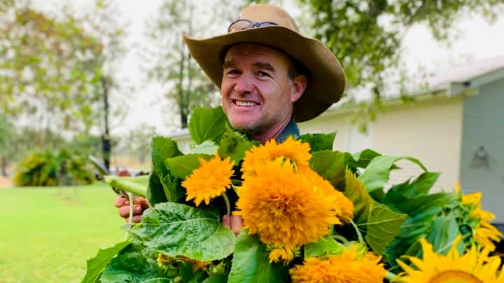 A man wearing a wide-brimmed hat smiles while holding sunflowers.