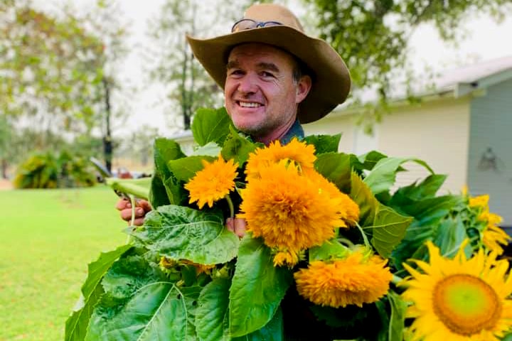 A man wearing a wide-brimmed hat smiles while holding sunflowers.