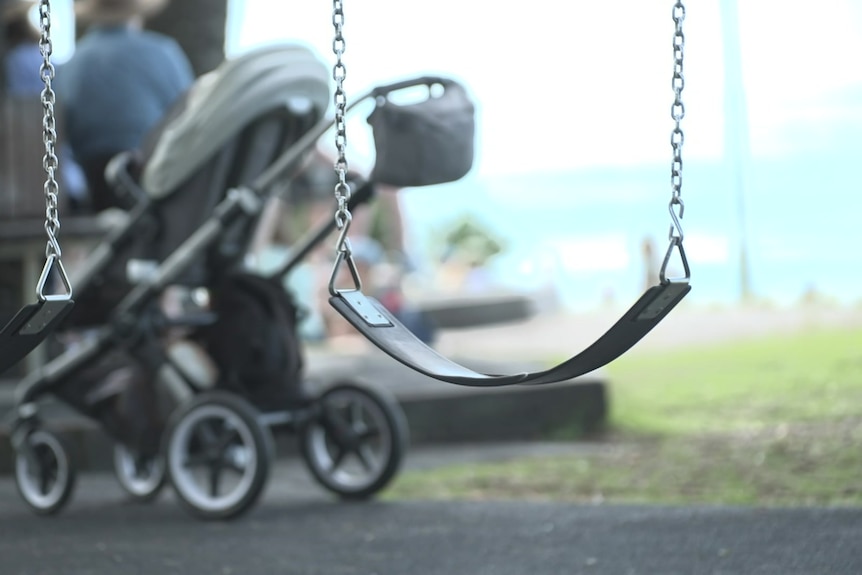 Empty swings in a park with a pram parked in the background.