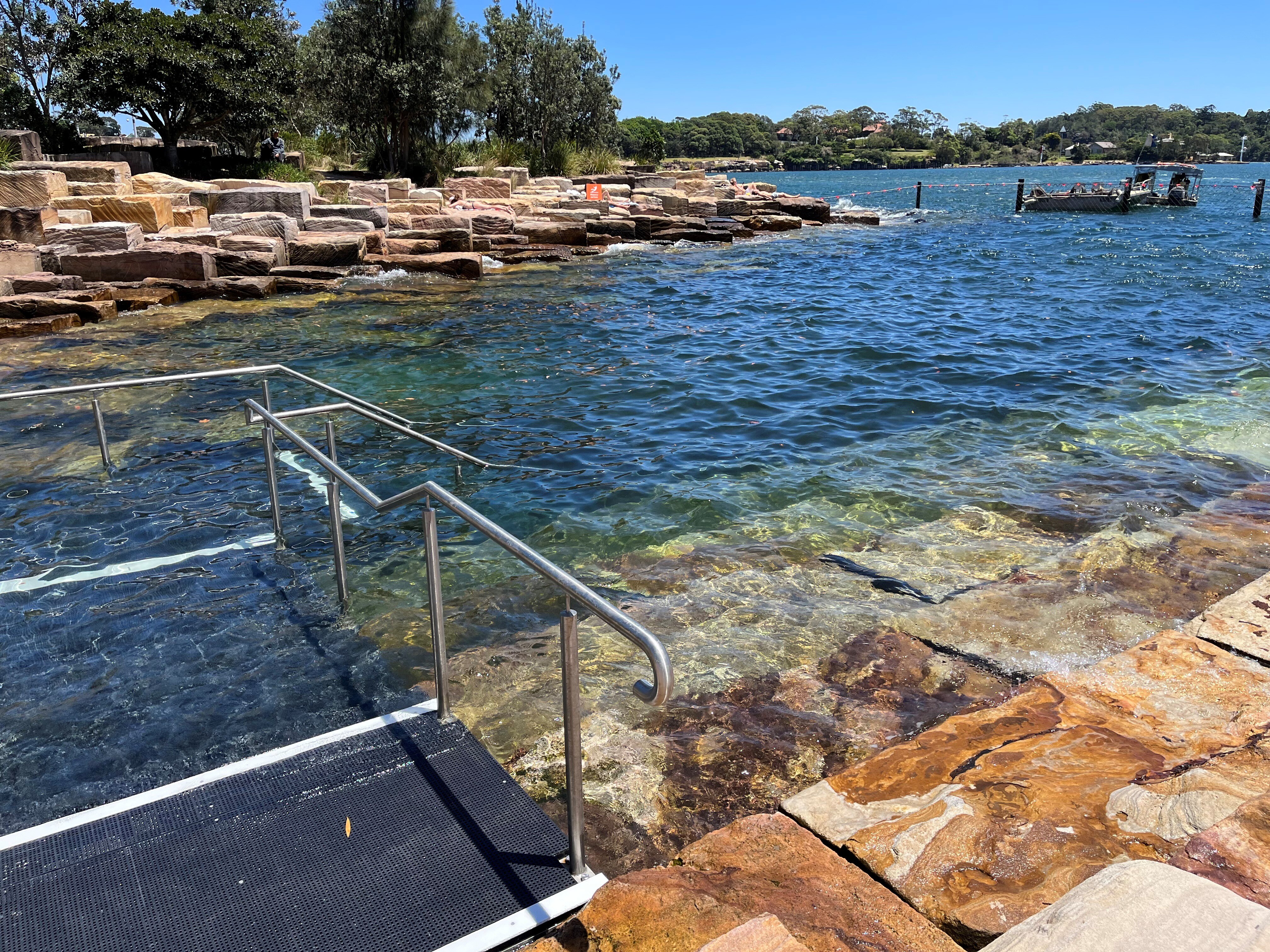 A black accessible ramp with railings leading down some large rocks into a swimming area.