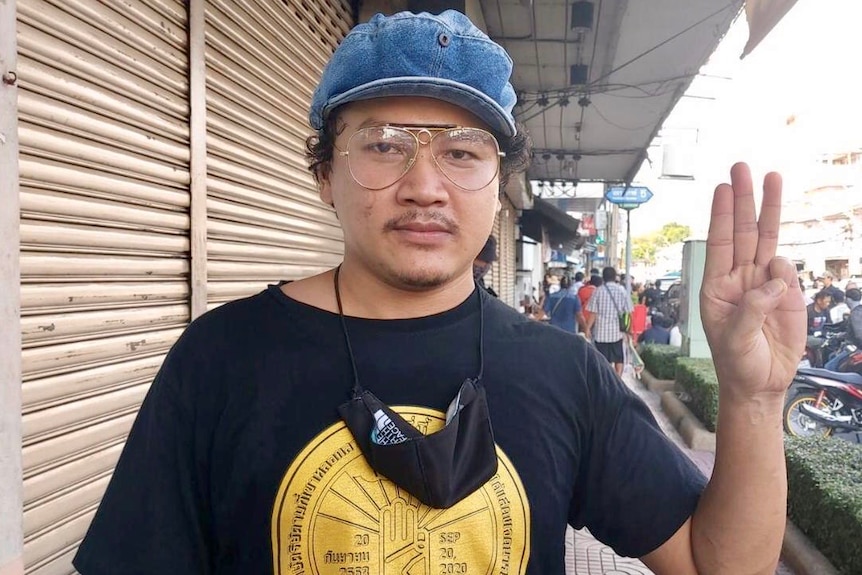 Man wearing large glasses and blue hat holds up three fingers on left hand