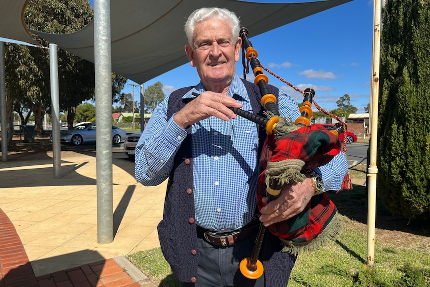 A white-haired man wearing a blue checked shirt and blue vest jacket holds bagpipes