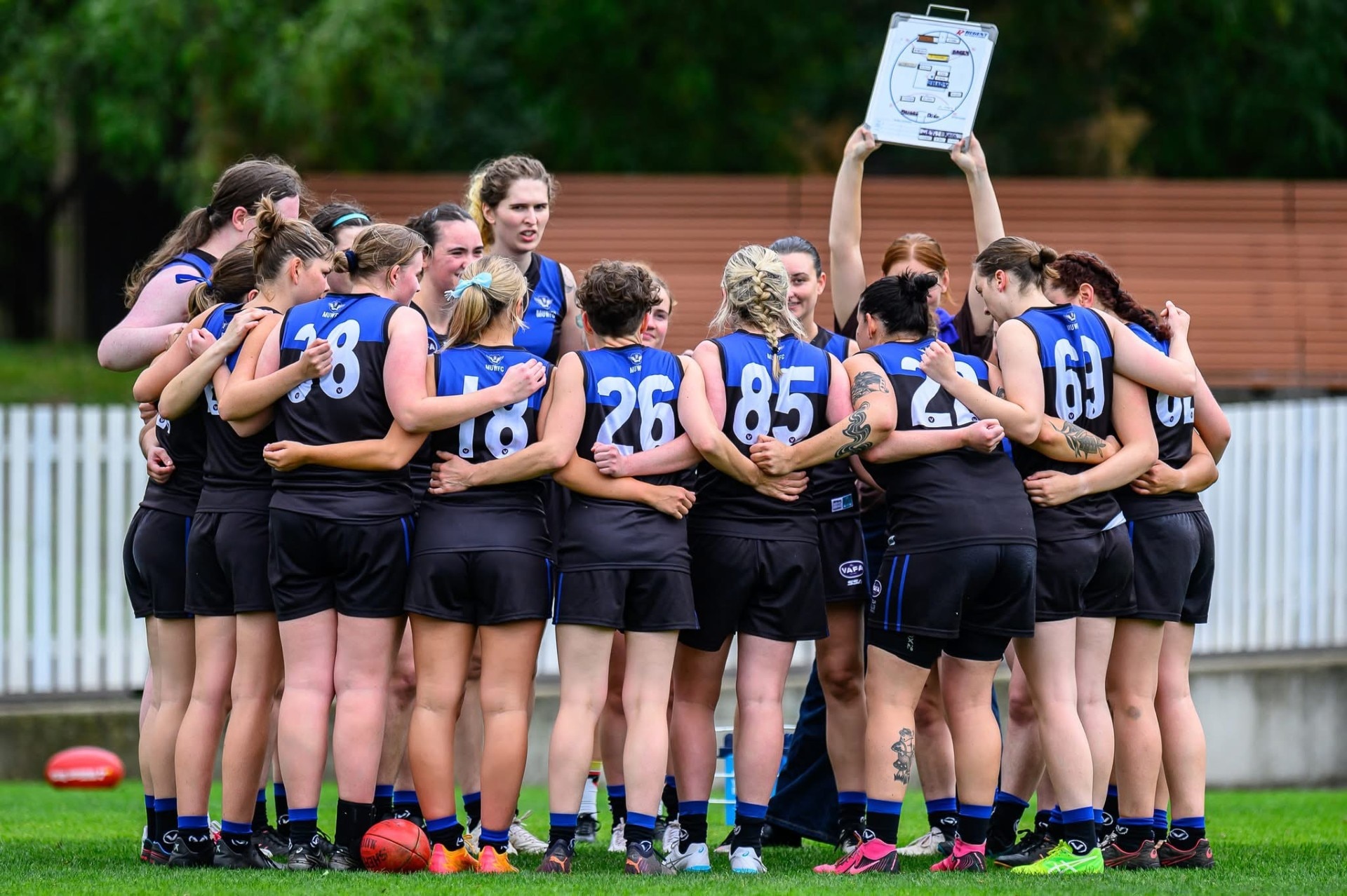 Um grupo de jogadores das Regras Aussie se reúne enquanto alguém segura um quadro branco com instruções.