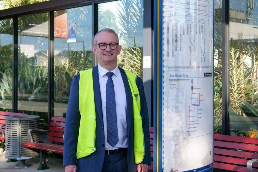 Grey haired man in black glasses, suit and high vis vest.