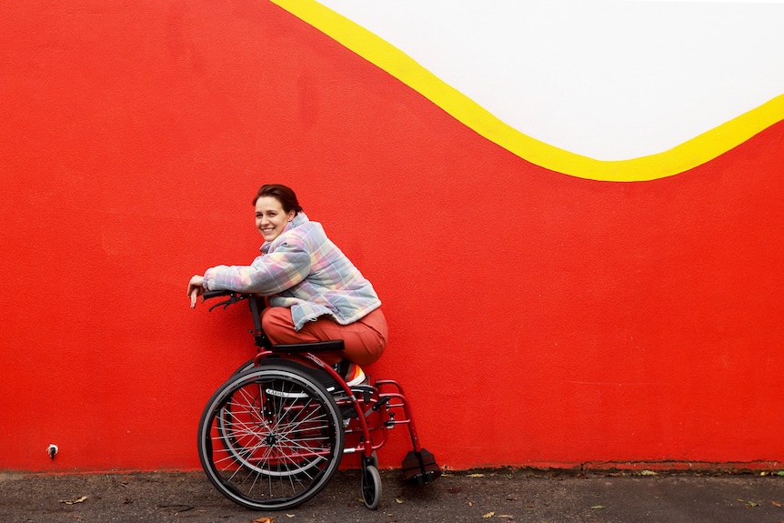 Tessa crouches atop her wheelchair and looks to the camera with a big grin, while positioned in front of a red and white wall.
