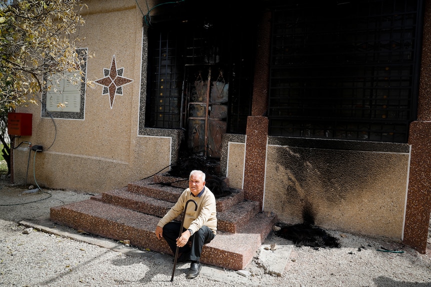 An older man with a walking stick sits on the front step of a house that is black, burned out by fire