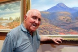 An elderly man stands in front of a landscape painting of a mountain range and kangaroos.