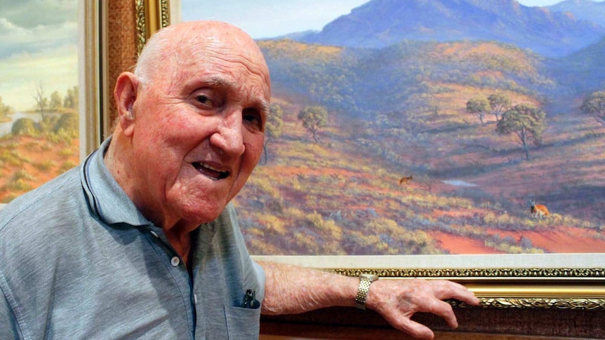 An elderly man stands in front of a landscape painting of a mountain range and kangaroos.