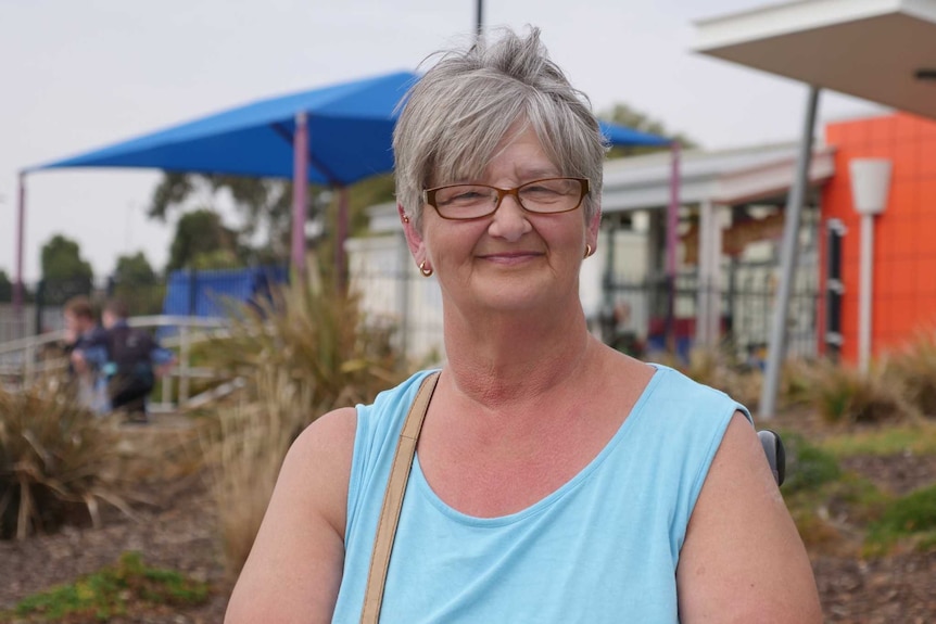 A woman with grey hair and glasses sits in a wheelchair, looking at the camera.