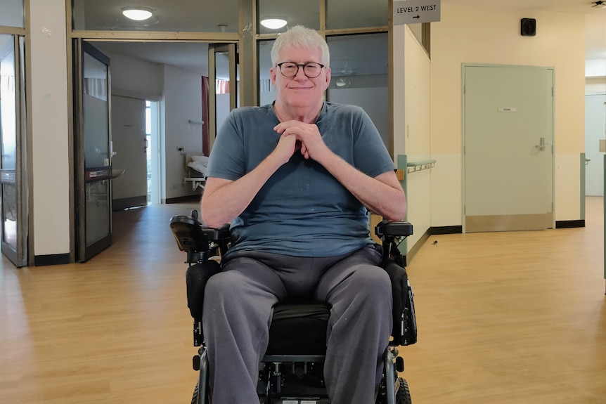 Michael Sibly wearing glasses and sitting in a wheelchair with his hands clasped together underneath his chin