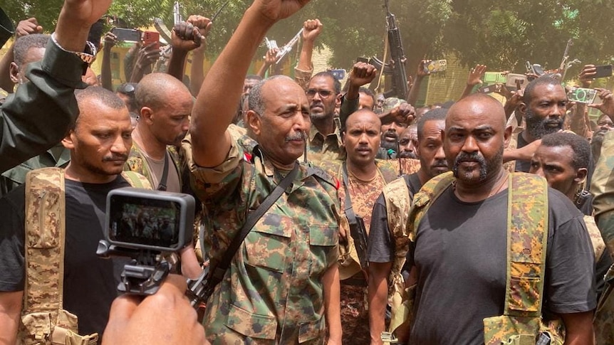 A group of Sudanese men in military gear stand in a group with arms raised in the air. 