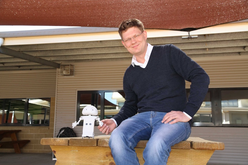 Academic Simon Egerton sits on a table in a school yard. He reaches down to hold the hand of a robot he's designed.