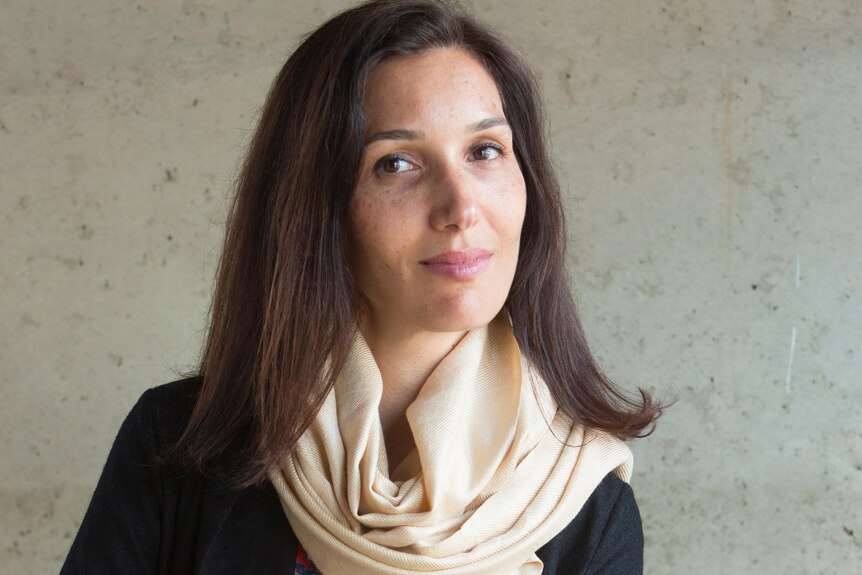 Delphine Minoui, with shoulder-length brown hair, scarf and slight smile, sits on a chair with arms crossed over knee.