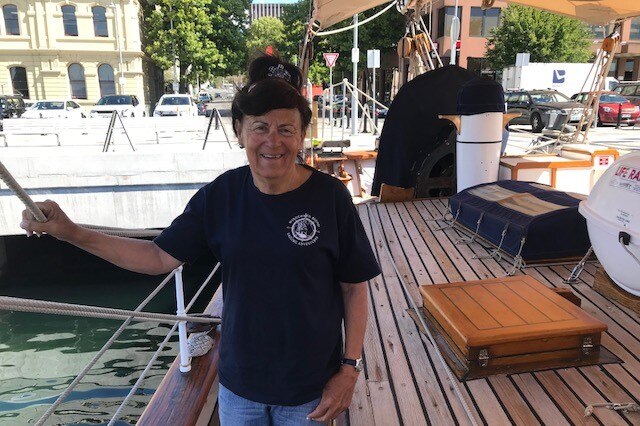 A woman in a black t-shirt stands smiling on the wooden deck of a large boat.