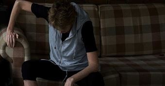 A woman steadies herself on the armrest of a sofa while sitting and looking at the floor in pain.