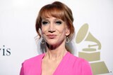 A portrait shot of Kathy Griffin standing in front of a backdrop showing the Grammys logo.