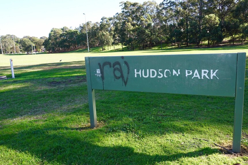A graffitied green Hudson Park sign in front of park.