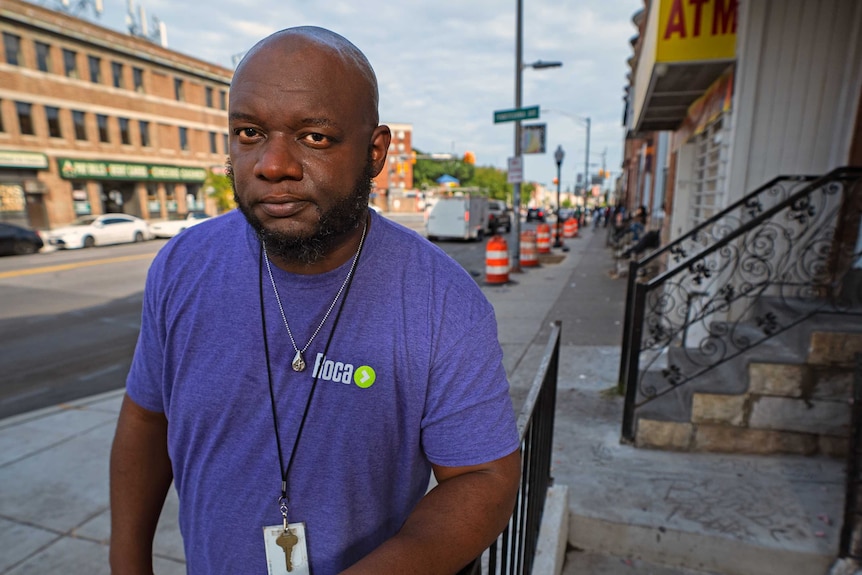 A man in a blue t-shirt leaning against a fence railing on a Baltimore street