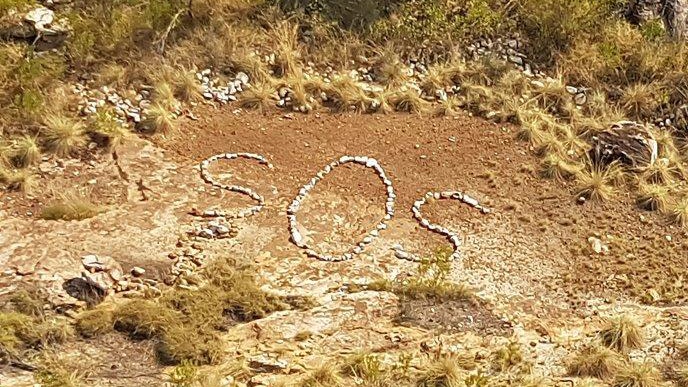 SOS sign picked out in rocks on scrubby ground.