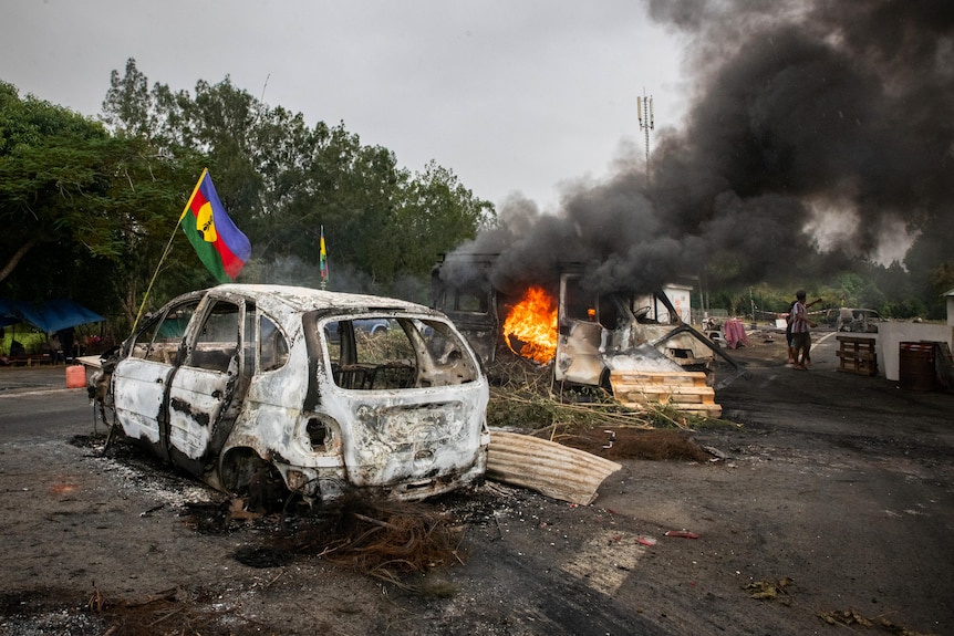 A burnt-out car sits next to a burning car with flames and black smoke pouring from it on a motorway surrounded by green trees