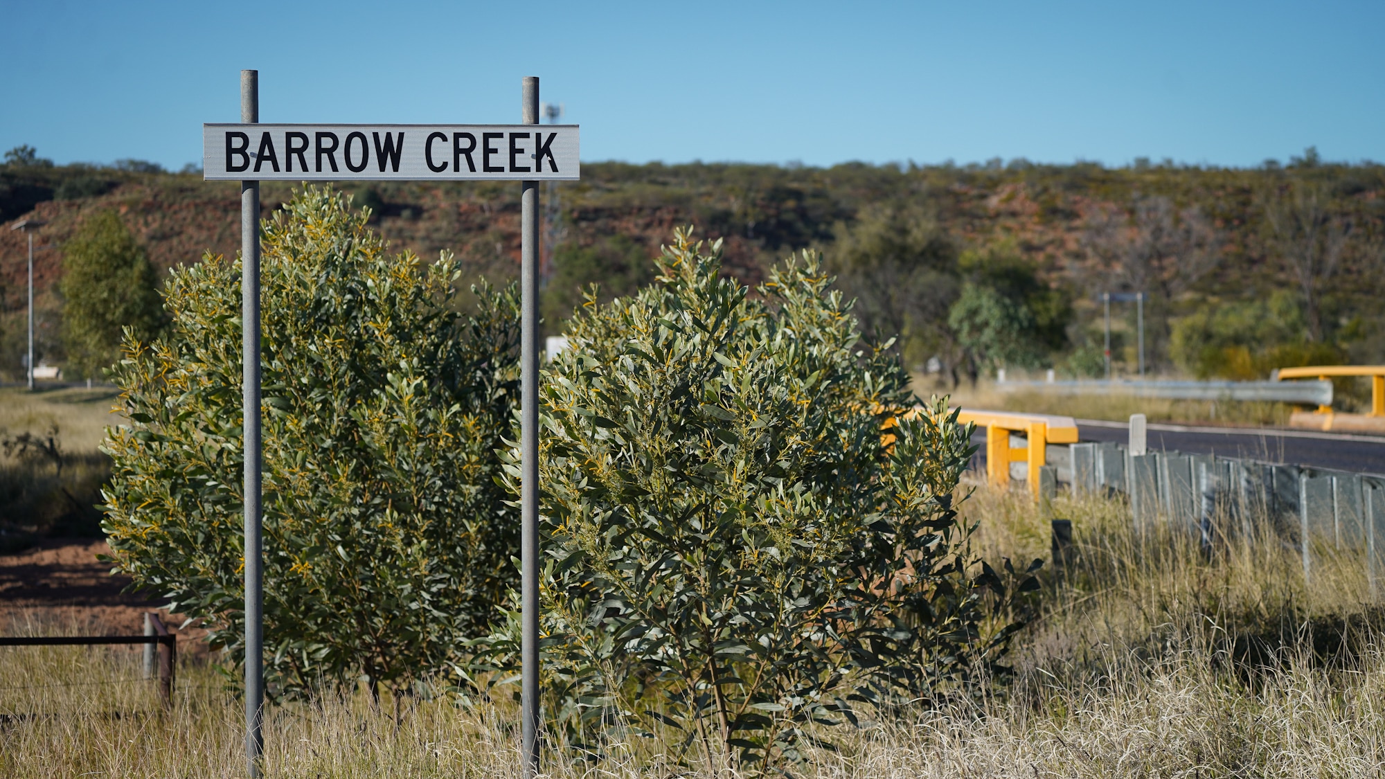 An outback landscape runs along a highway, a roadside sign in the tall grass reads: BARROW CREEK
