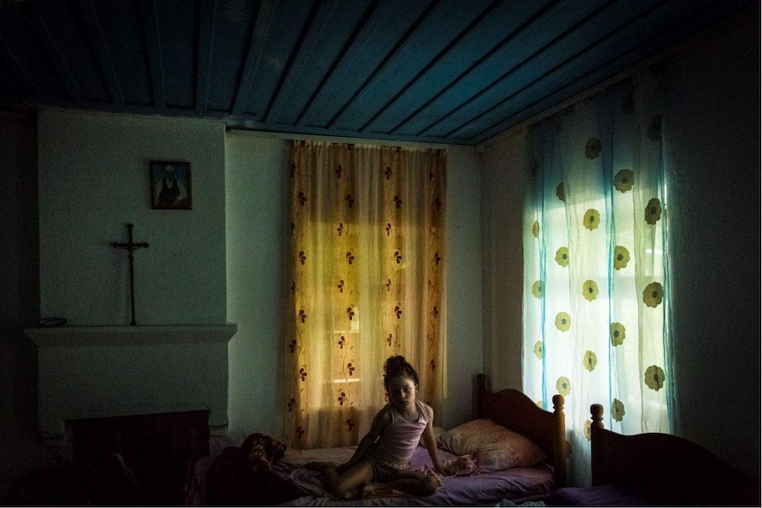 Young girl sitting up in bed in a dark room with light coming through window with floral curtains