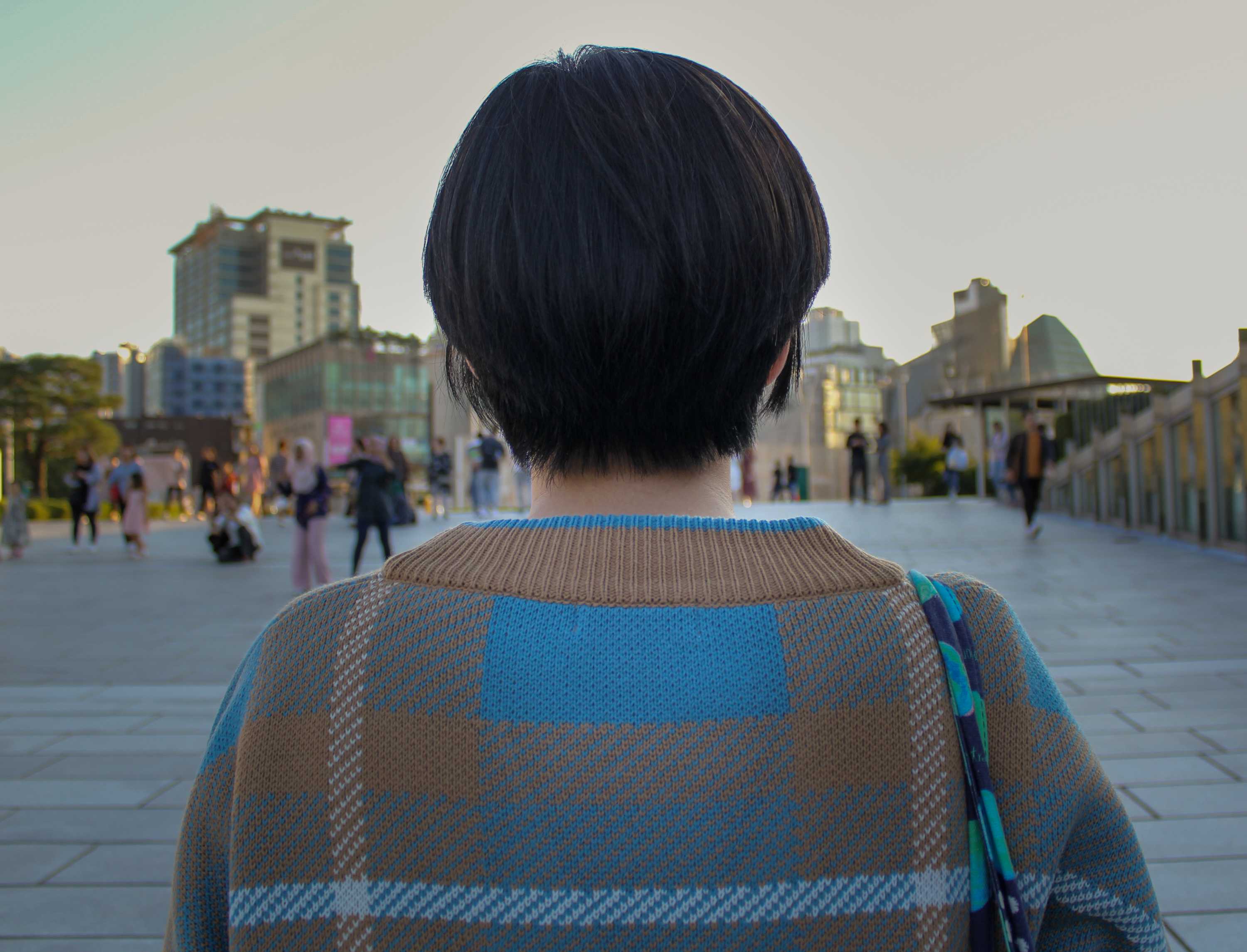 A woman with a short black bob haircut with her back to the camera with a university campus in the background.