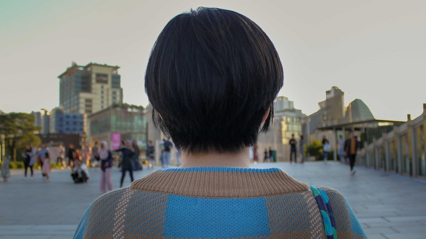 A woman with a short black bob haircut with her back to the camera with a university campus in the background.
