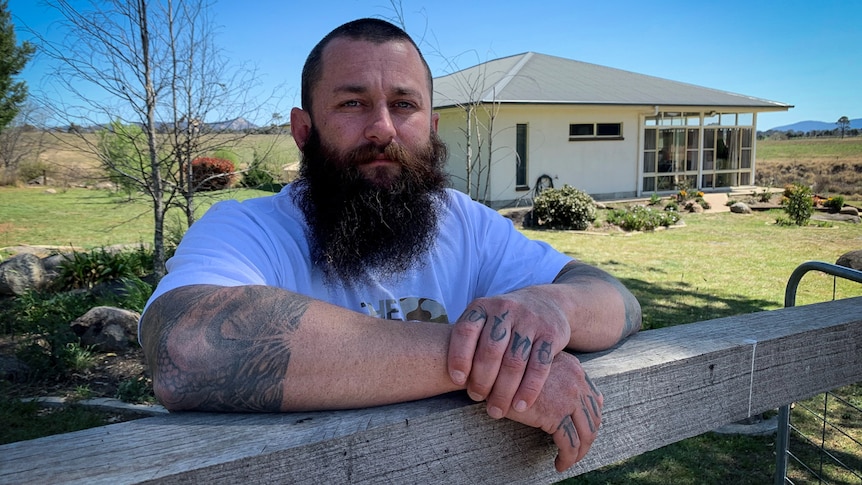 A man with a beard and tattoos leans on a fence in front of a rural property.