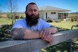 A man with a beard and tattoos leans on a fence in front of a rural property.