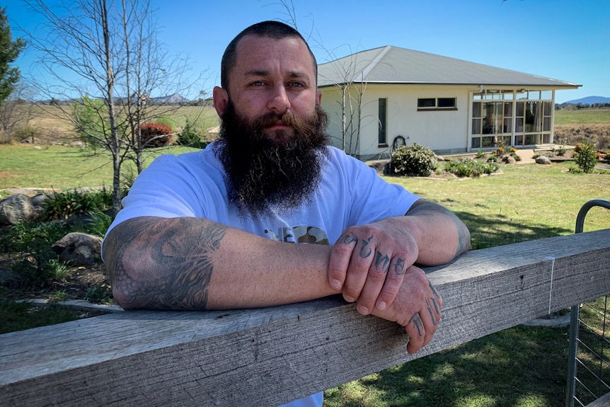 A man with a beard and tattoos leans on a fence in front of a rural property.