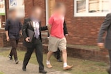 A photo of four men with their faces blurred, three of them in suits leading away a man in a red shirt in handcuffs.