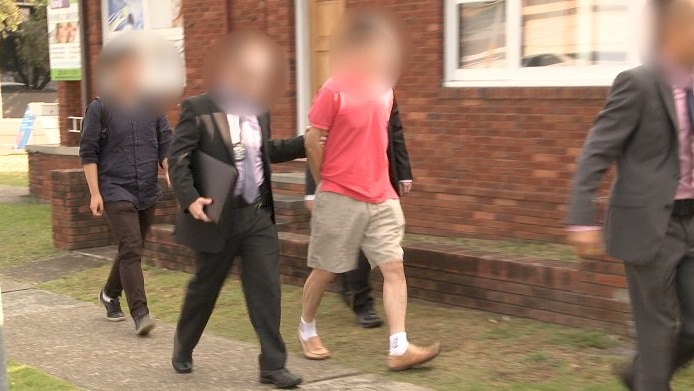 A photo of four men with their faces blurred, three of them in suits leading away a man in a red shirt in handcuffs.