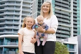 Stephanie holding her baby son Archie with her young daughter Lily by her side, standing in front of high rise apartments