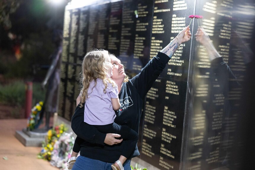 A woman holding a flower in her left hand while carrying her young daughter pays her respects at a memorial wall.   