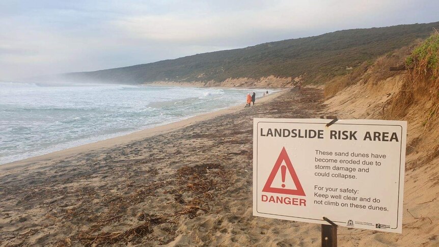 Waves washing up a beach with dunes in the background and sign warning of landslide risk from erosion and storms.