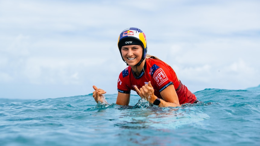 Molly Picklum gives shakas to the camera while on a surfboard in the ocean. She is wearing a Red Bull helmet.