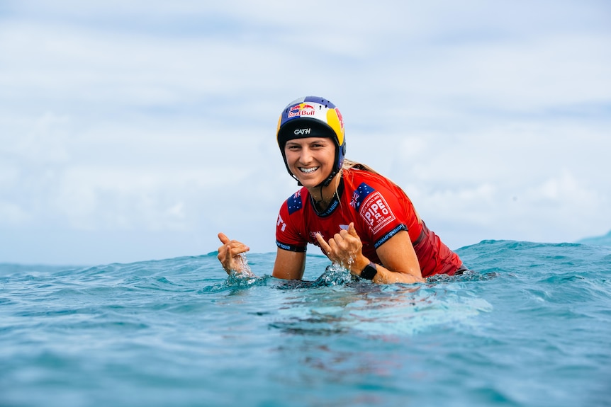 Molly Picklum gives shakas to the camera while on a surfboard in the ocean. She is wearing a Red Bull helmet.