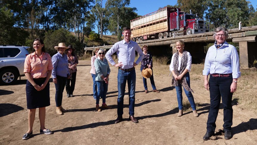 A group of men and women stand apart in a clearing below an old wooden bridge. A cattle truck passes on the bridge behind them.