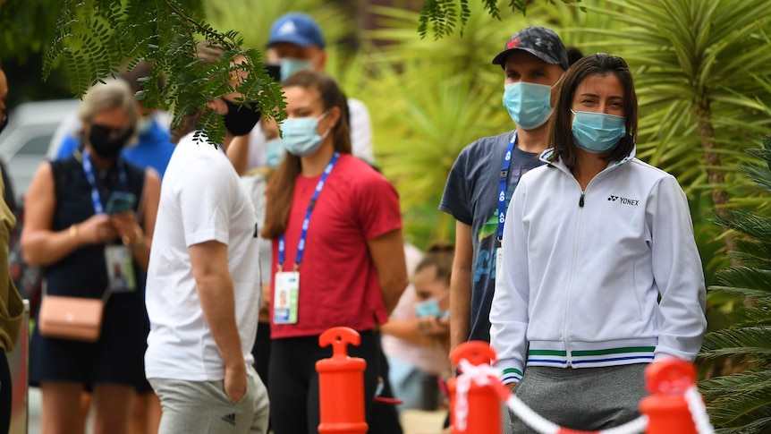 Tennis players wearing masks line up for COVID-19 tests outside, surrounded by plants.