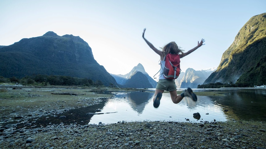 Woman jumps for joy in front of a picturesque lake in New Zealand which is surrounded by mountains.