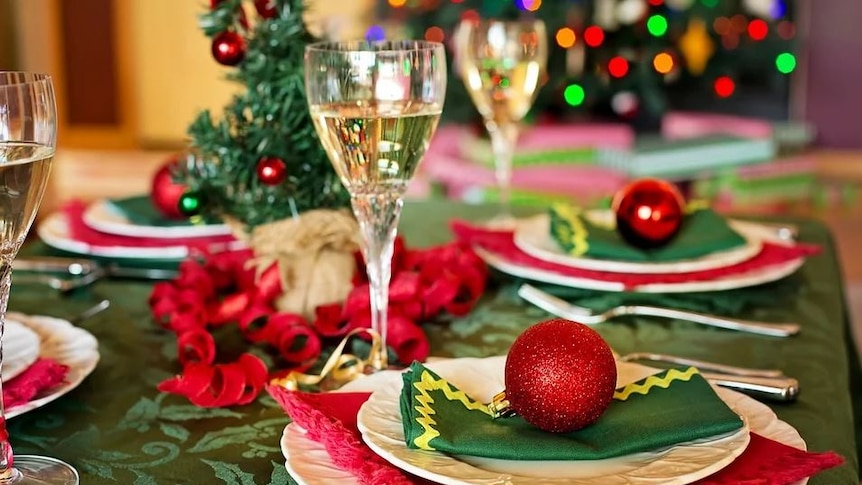 Table with Christmas decorations, plates, baubles, glasses of wine, Christmas tree in background.
