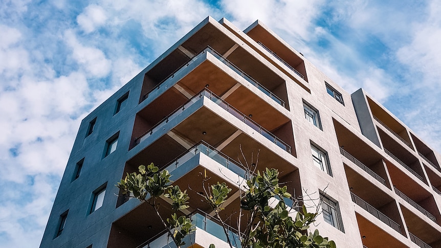 From the ground up, a large brown brick apartment complex stands, with a cloudy blue sky backdrop.