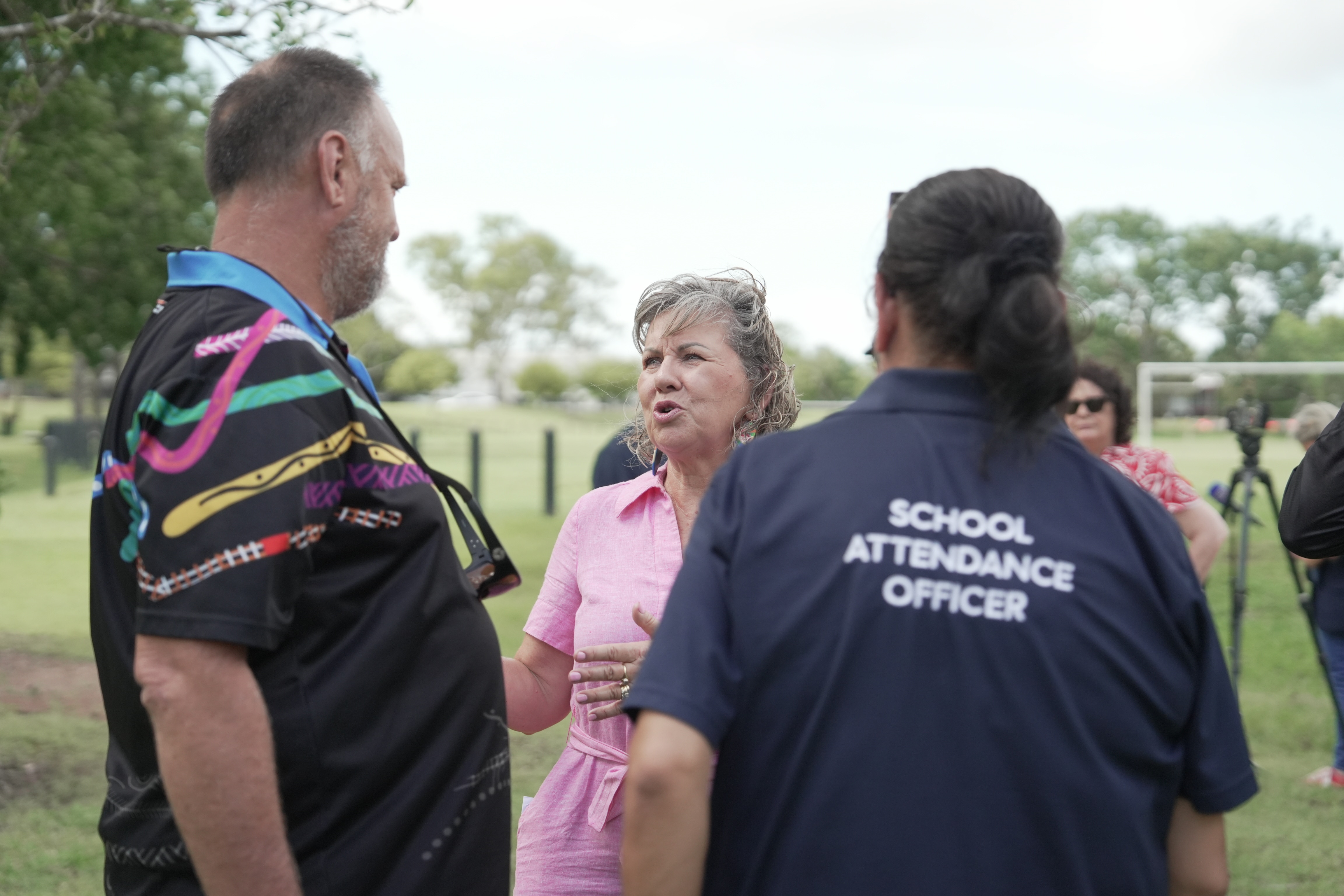 Uma mulher de vestido rosa conversando com as pessoas, uma delas usando uma camiseta que diz: OFICIAL DE ATENDIMENTO ESCOLAR