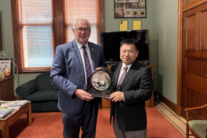 Two men in suits stand in an office holding a clock-like ornament featuring pictures of pandas.