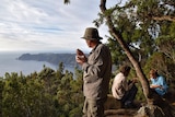 A bushwalker on the Three Capes Track in Tasmania looks out at the bush view from the top of Mount Fortescue while drinking tea.
