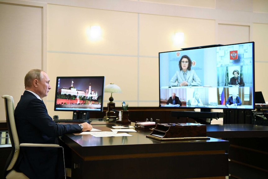 An elderly man in a dark suit sits at a desk and faces a bank of video conference monitors.